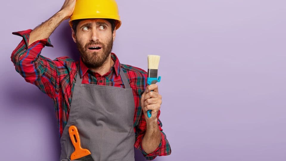 Nervous construction worker holds paint brush, touches head and looks aside, wears safety hardhat, dressed in apron, puzzled with having much work, poses on purple wall with blank copy space