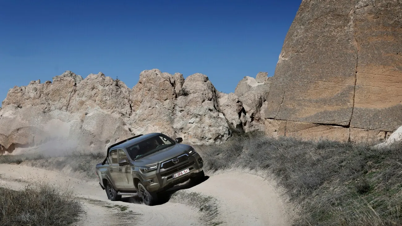 Der Hilux Hybrid fährt eine Kurve auf einem Weg neben Hügeln.