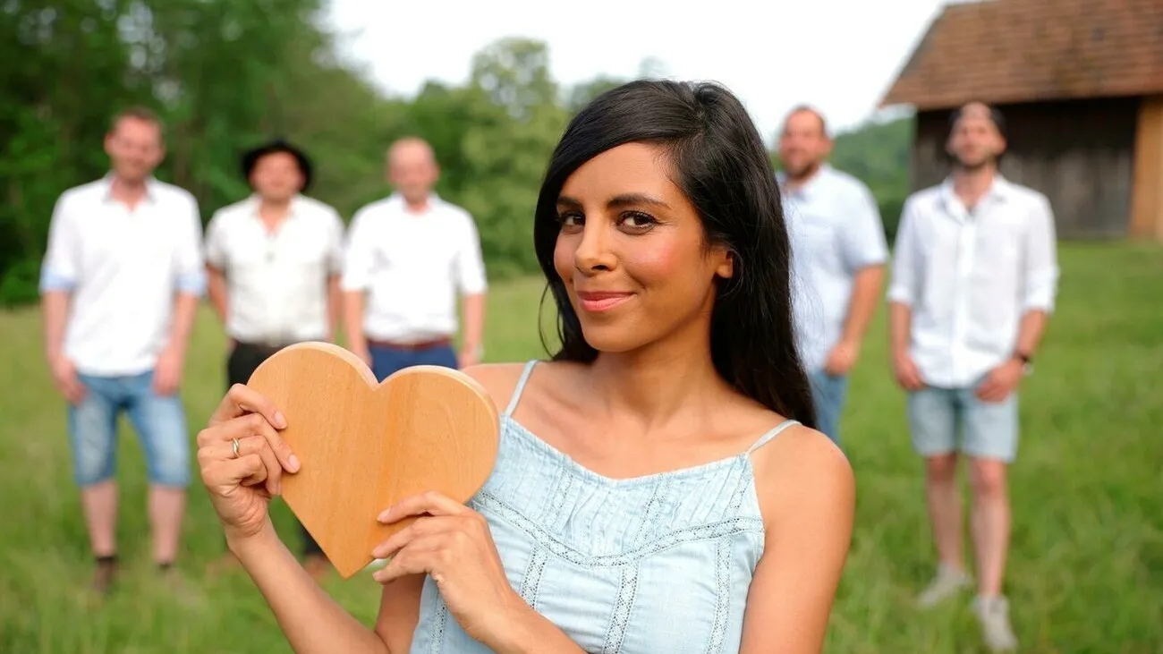 Collien Ulmen-Fernandes steht mit einem Holzherz in den Händen vorne im Bild und lächelt. Im Hintergrund sind die fünf Männer der Show.