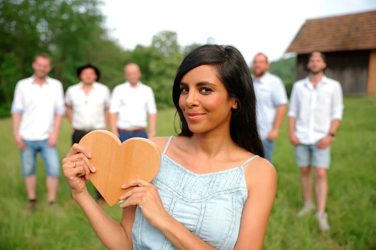 Collien Ulmen-Fernandes steht mit einem Holzherz in den Händen vorne im Bild und lächelt. Im Hintergrund sind die fünf Männer der Show.