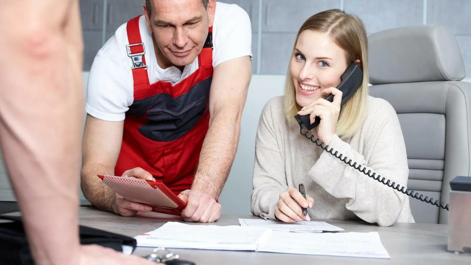 Eine Frau sitzt an einem Tisch, lächelt und telefoniert, neben ihr steht ein Handwerker mit einer roten Latzhose und zeigt auf ein Infobrett in seinen Händen.