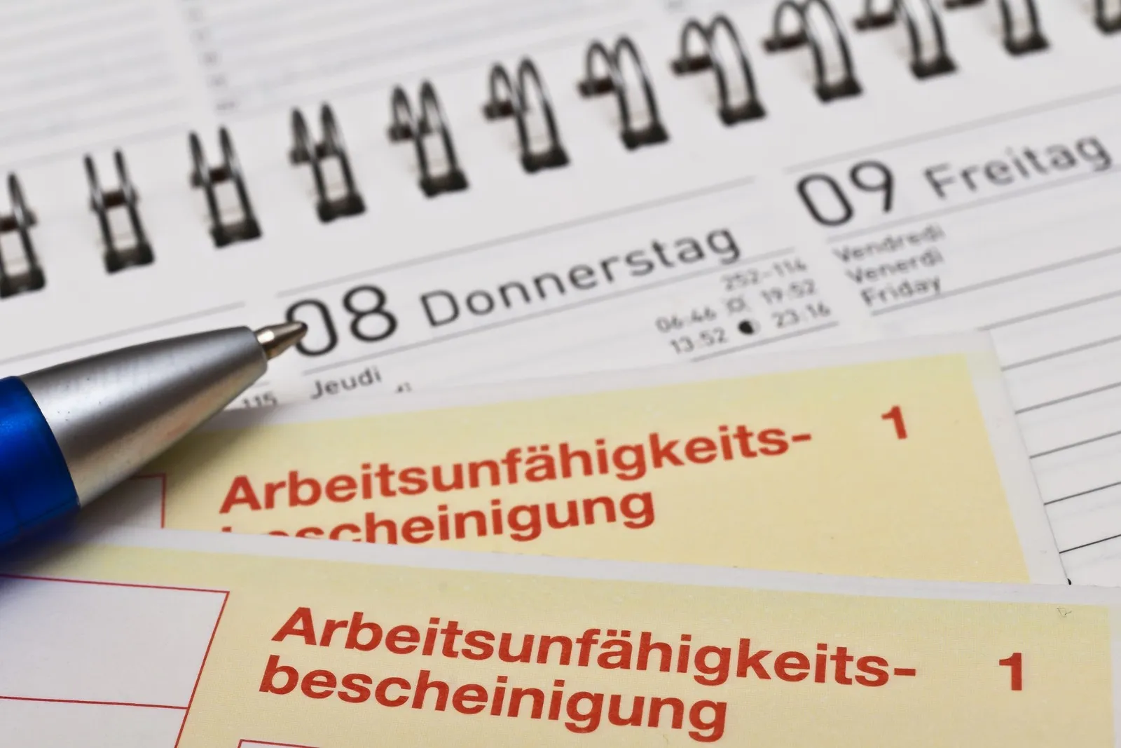 Zwei Krankenscheine liegen auf einem Kalender, von links ist der obere Teil eines blauen Kugelschreibers zu sehen.