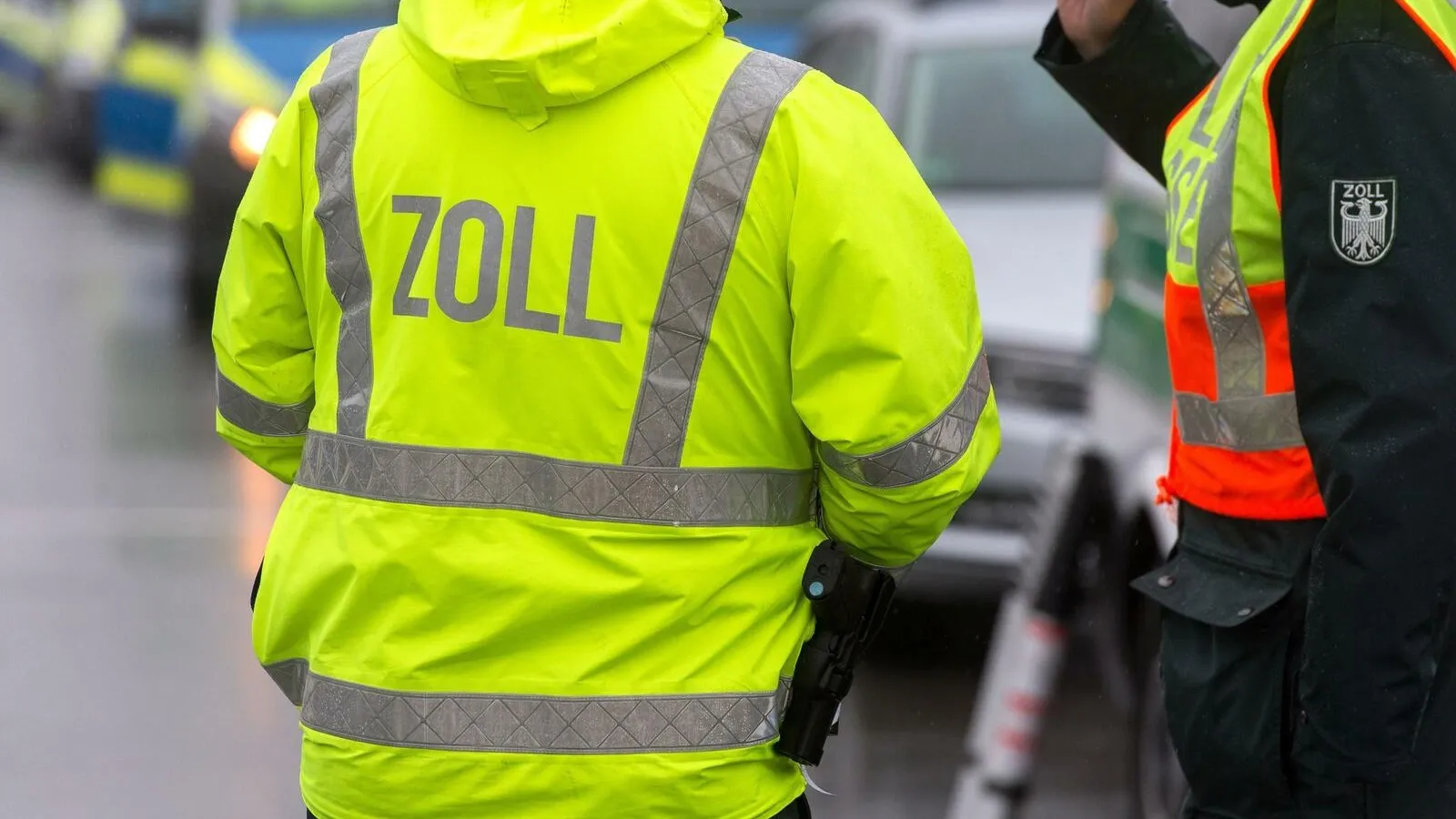 Ein Mann in einen gelben Jacke steht mit dem Rücken zum Bild. Es ist die Aufschrift "ZOLL" zu erkennen.