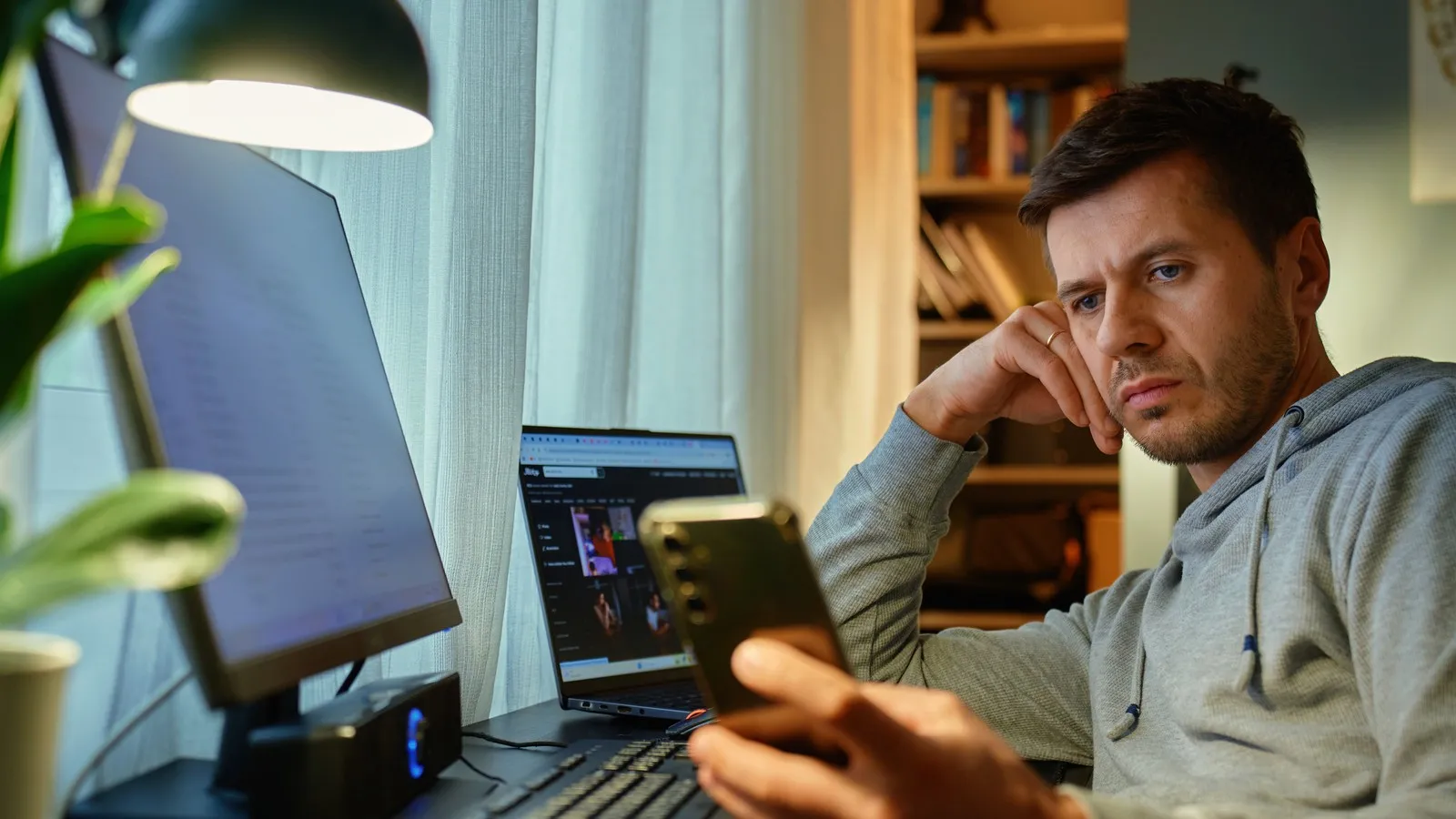 Ein Mann schaut gelangweilt auf sein Smartphone. Vor ihm auf dem Schreibtisch stehen Laptop und ein Computer.