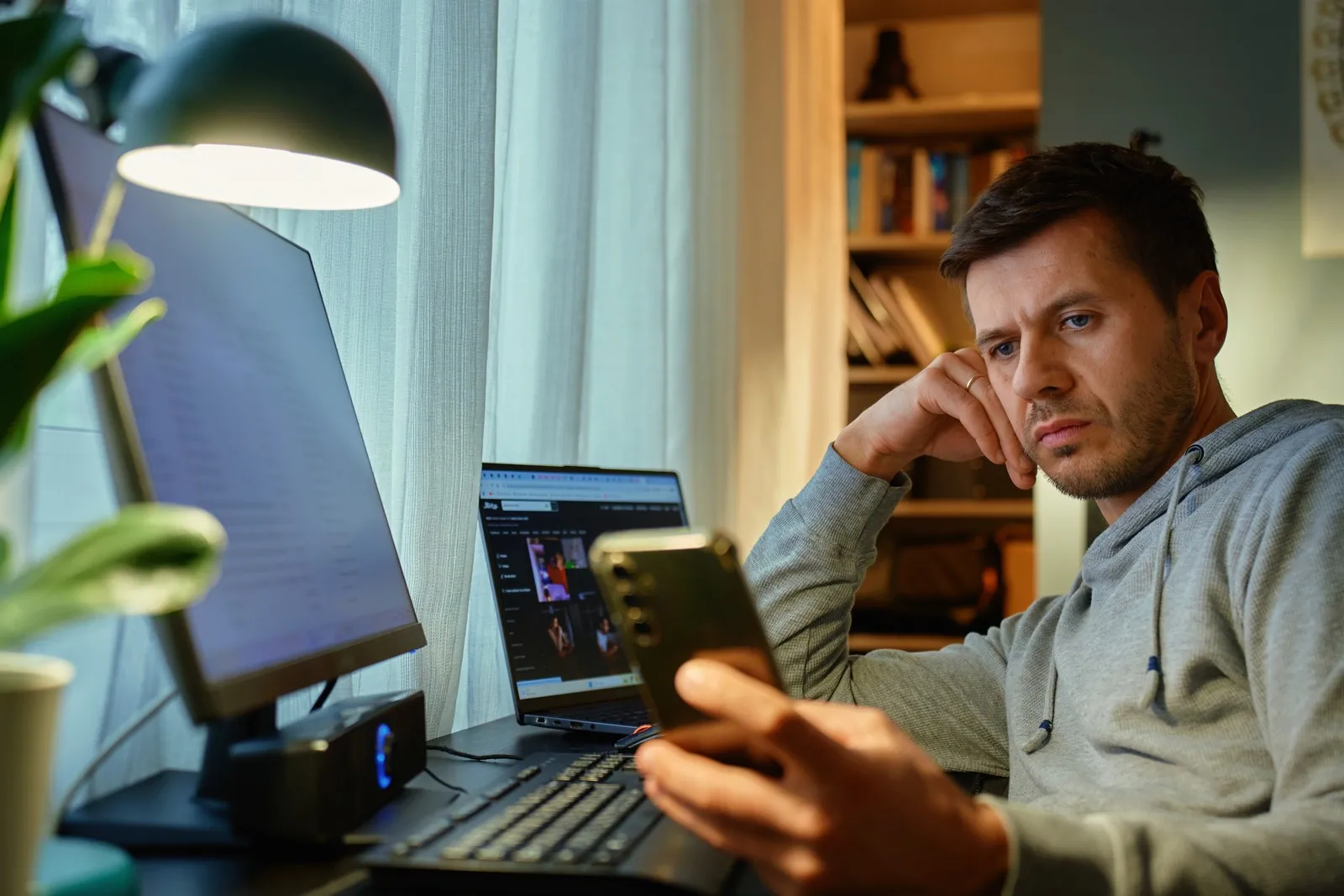 Ein Mann schaut gelangweilt auf sein Smartphone. Vor ihm auf dem Schreibtisch stehen Laptop und ein Computer.