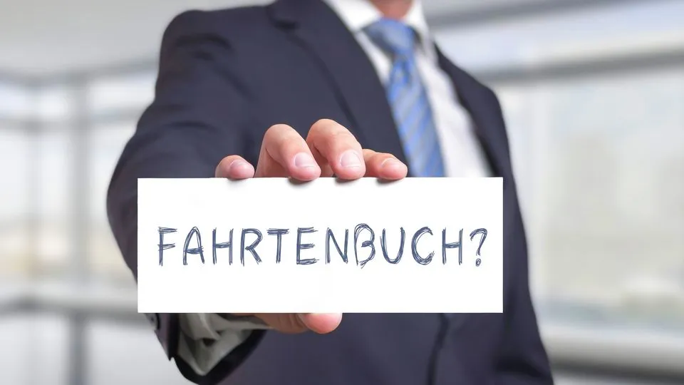 Eine Person im Anzug hält ein kleines weißes Schild hoch, auf dem "Fahrtenbuch?" steht.