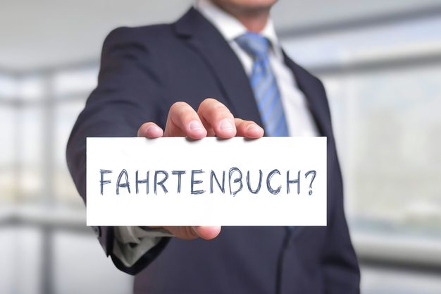 Eine Person im Anzug hält ein kleines weißes Schild hoch, auf dem "Fahrtenbuch?" steht.