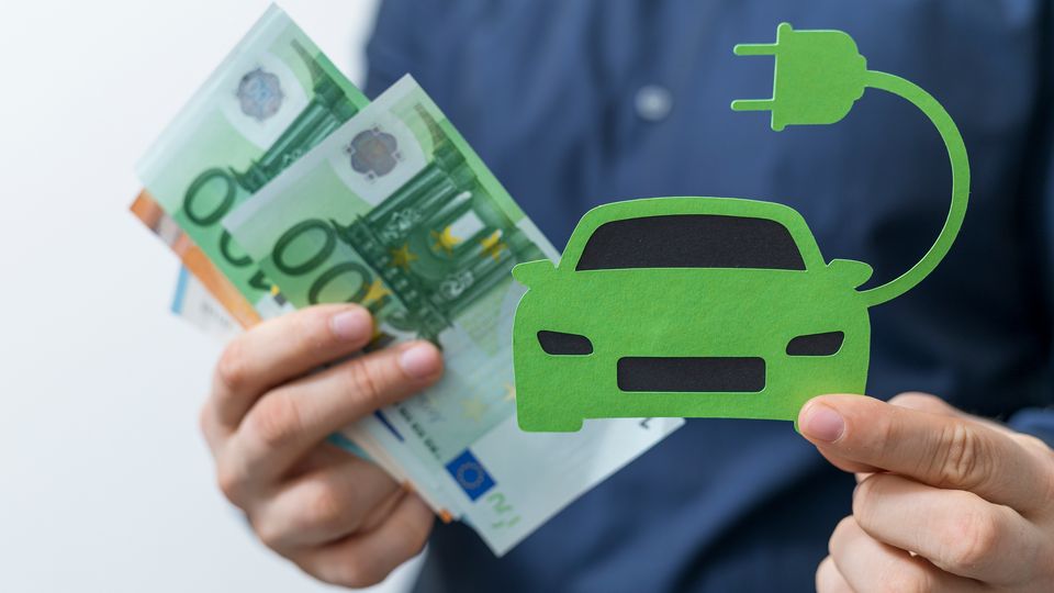 Mann hält ein E-Auto in der Hand und in der anderen Hand hat er mehrere Geldscheine