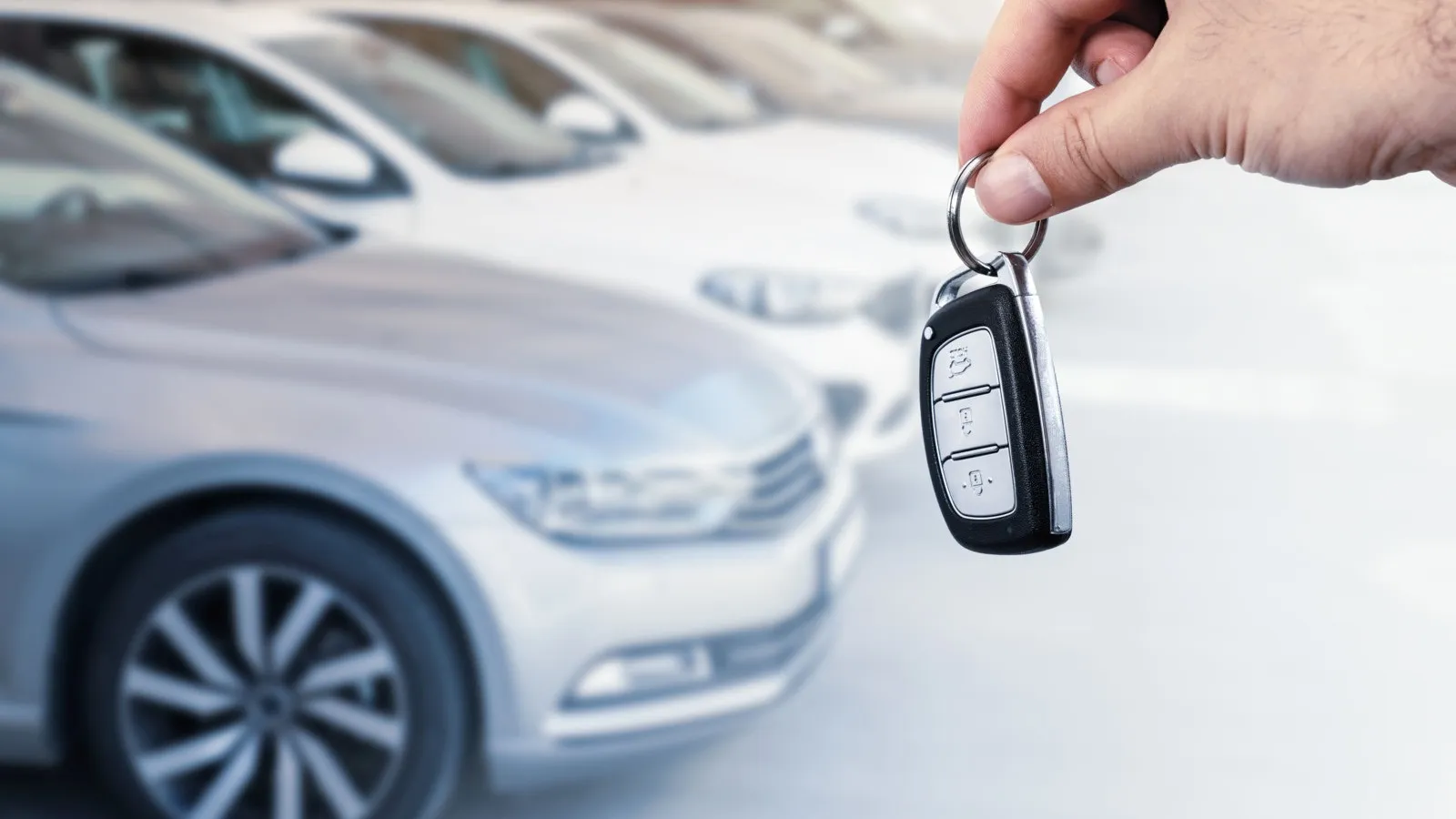 Eine Hand hält einen Autoschlüssel in der Luft. Im Hintergrund sind links Autos in einer Reihe zu sehen.