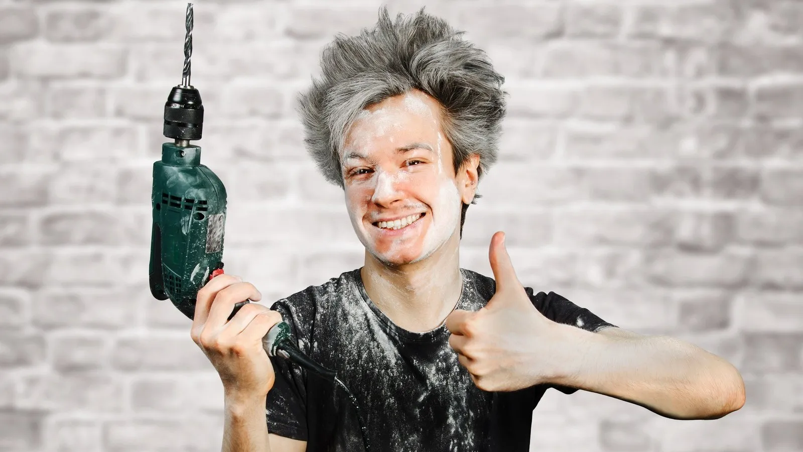 Ein schmutziger junger Handwerker mit Gipsflecken und einer Bohrmaschine in der Hand, zeigt den Daumen hoch und lächelt.