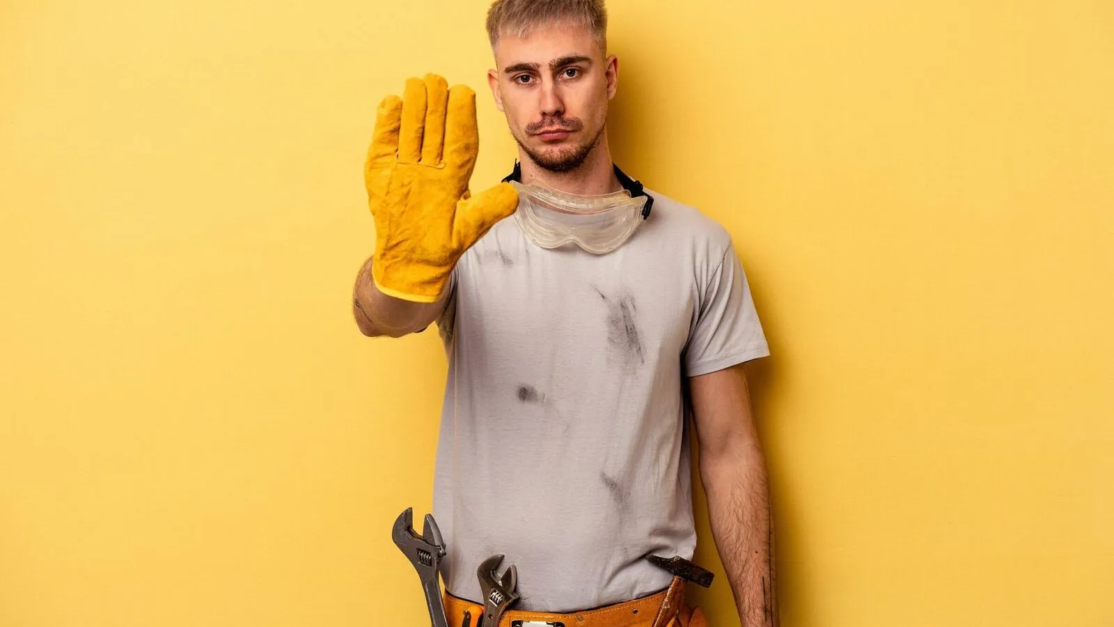 Ein Handwerker steht vor einem gelben Hintergrund und streckt seine Hand zu einem Stoppzeichen aus. Seine ausgestreckte Hand steckt in einem gelben Handschuh.