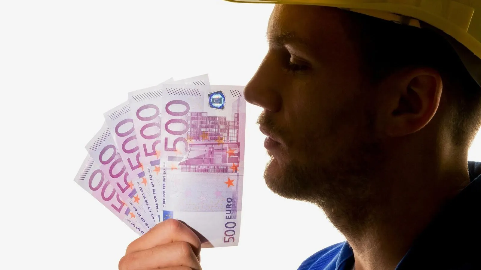 Das Gesicht eines Handwerkers, der einen gelben Schutzhelm trägt, ist von der Seite zu sehen. Er hält sich mehrere 500 Euro Scheine vor das Gesicht.