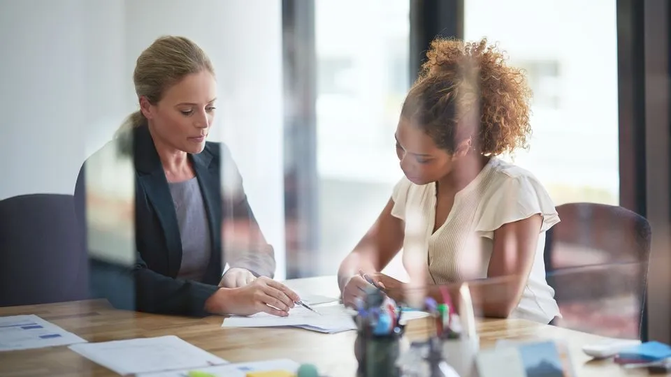 Zwei Frauen sitzen im Büro über ein Schriftstück gebeugt.