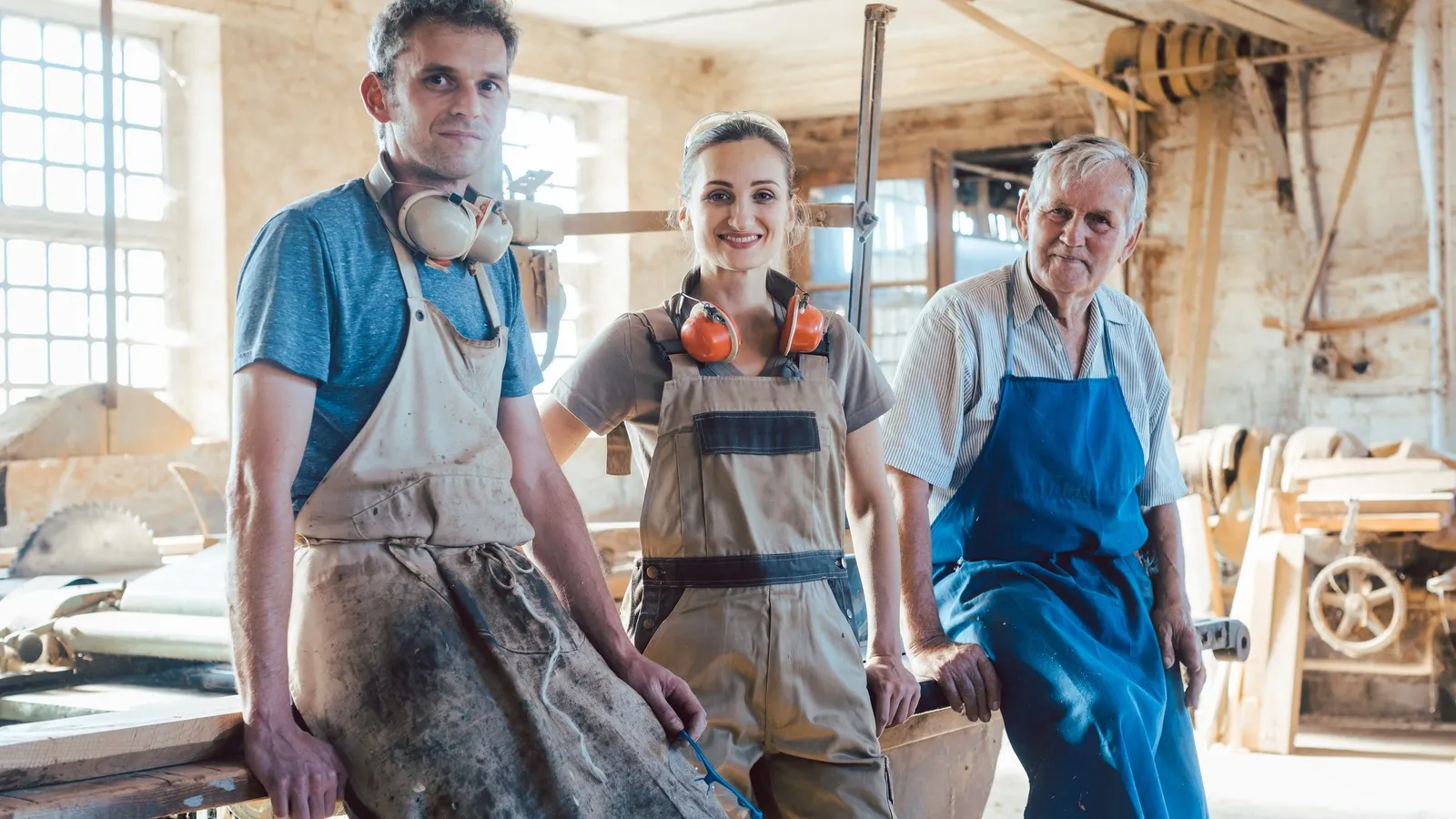 Eine Tischlerin und zwei Tischler stehen in der Werkstatt in Arbeitskleidung.