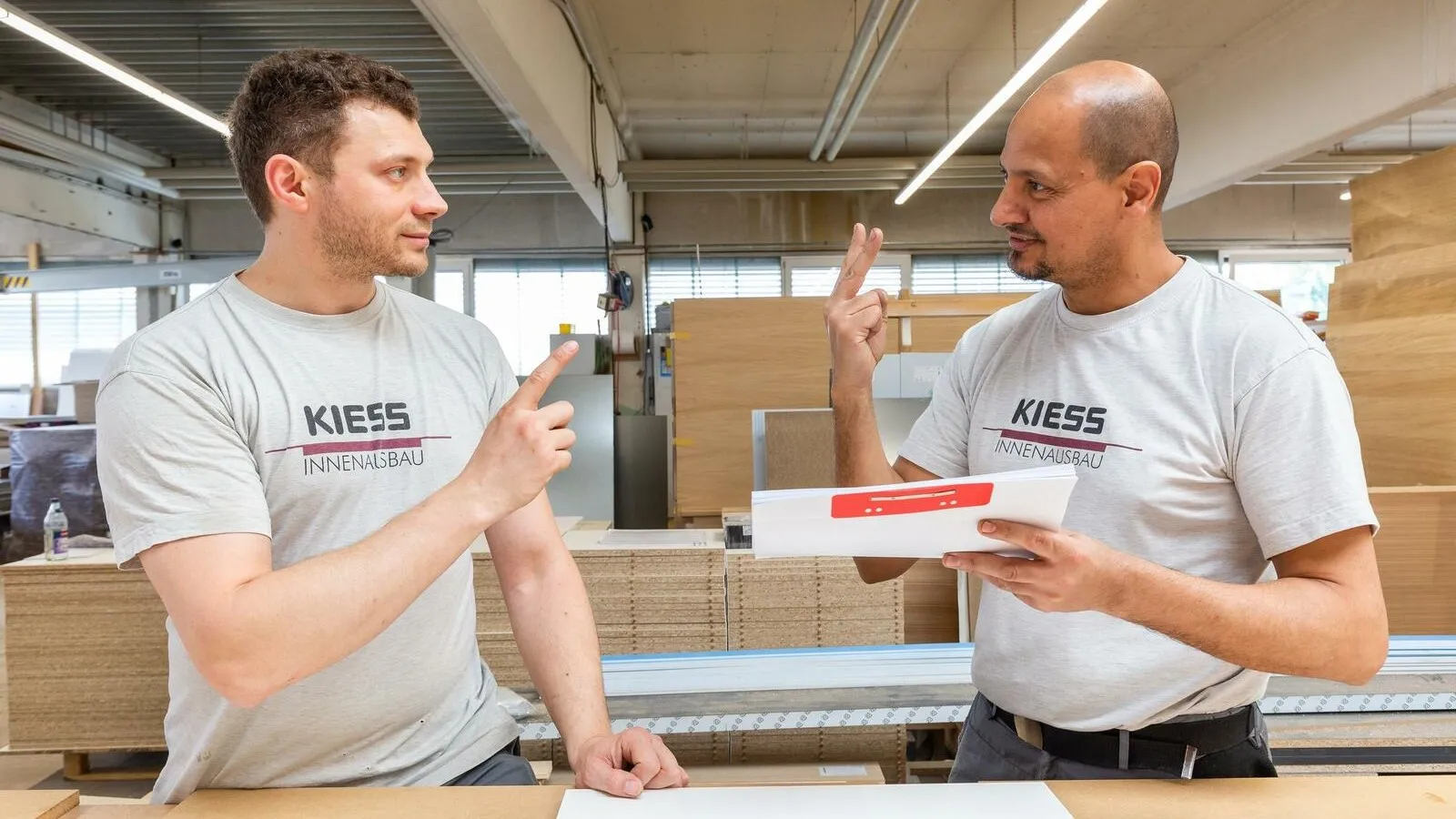 Zwei Mitarbeiter mit grauen T-Shirts und dem Firmenlogo der Alfred Kiess GmbH sprechen in Gebärdensprache miteinander im Lager.