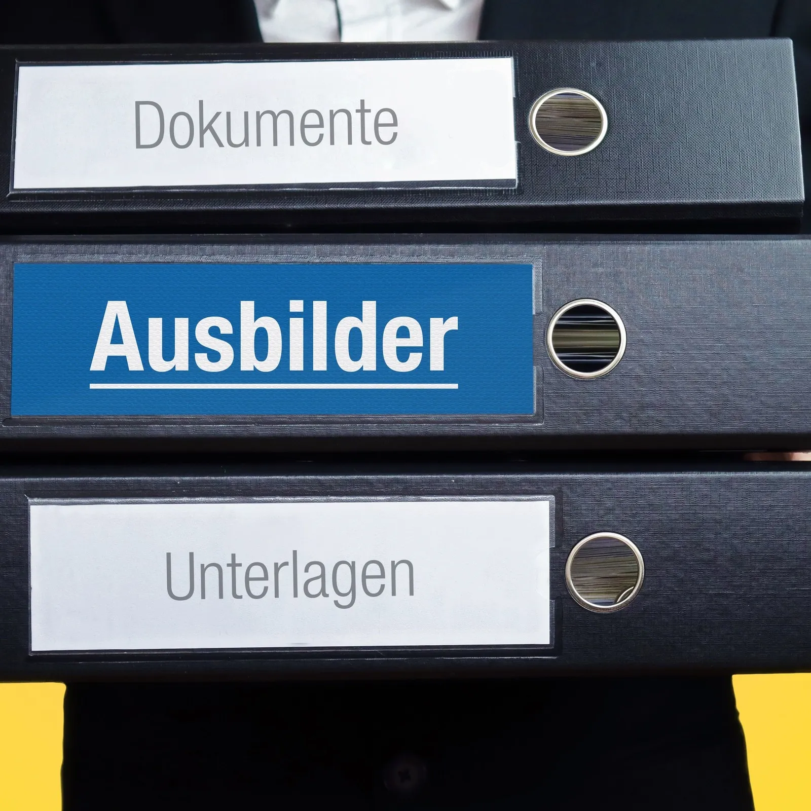 Ein Ausbilder trägt drei Ordner. SIe sind mit folgenden Wörtern beschriftet: Dokumente, Ausbilder, Unterlagen.