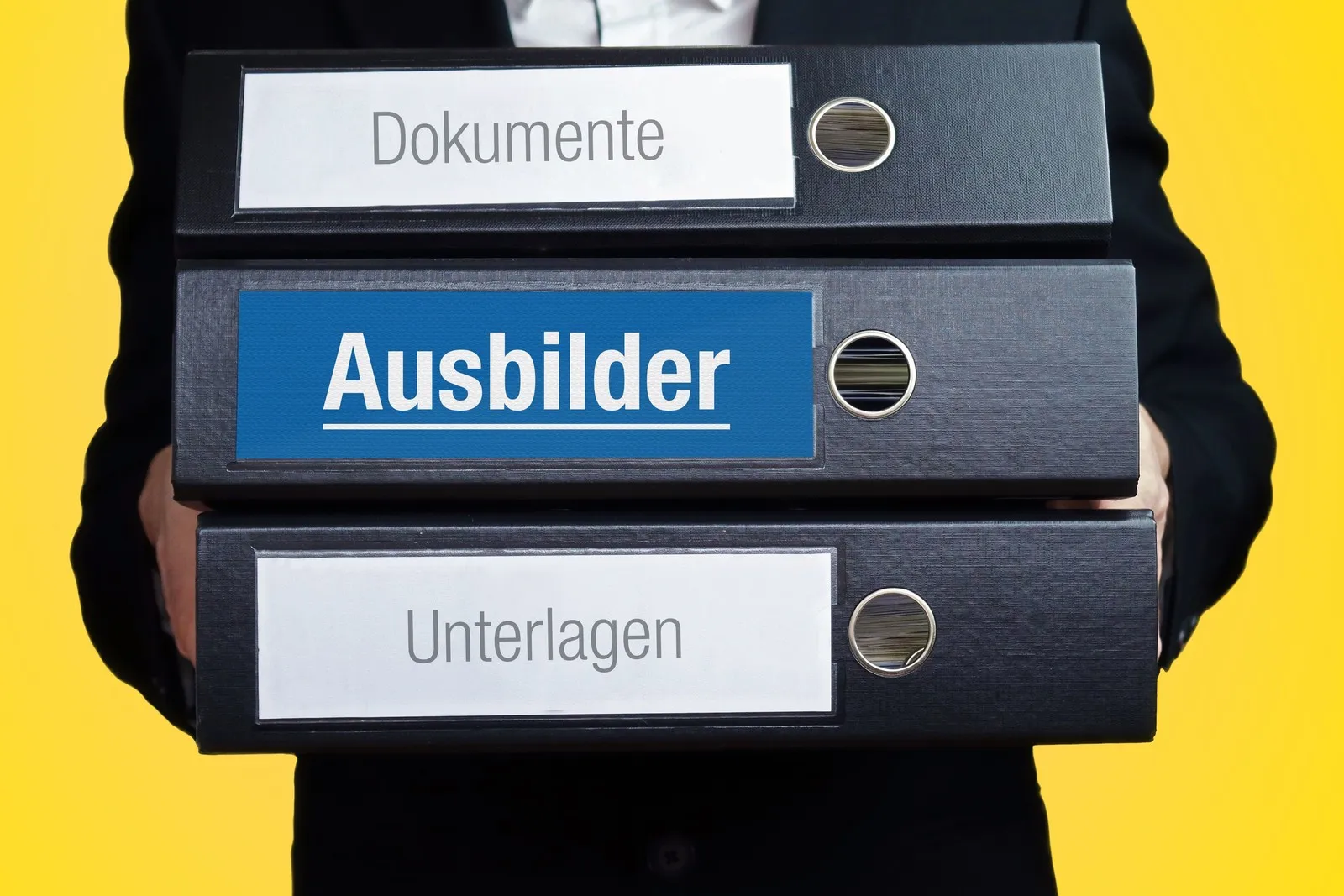 Ein Ausbilder trägt drei Ordner. SIe sind mit folgenden Wörtern beschriftet: Dokumente, Ausbilder, Unterlagen.