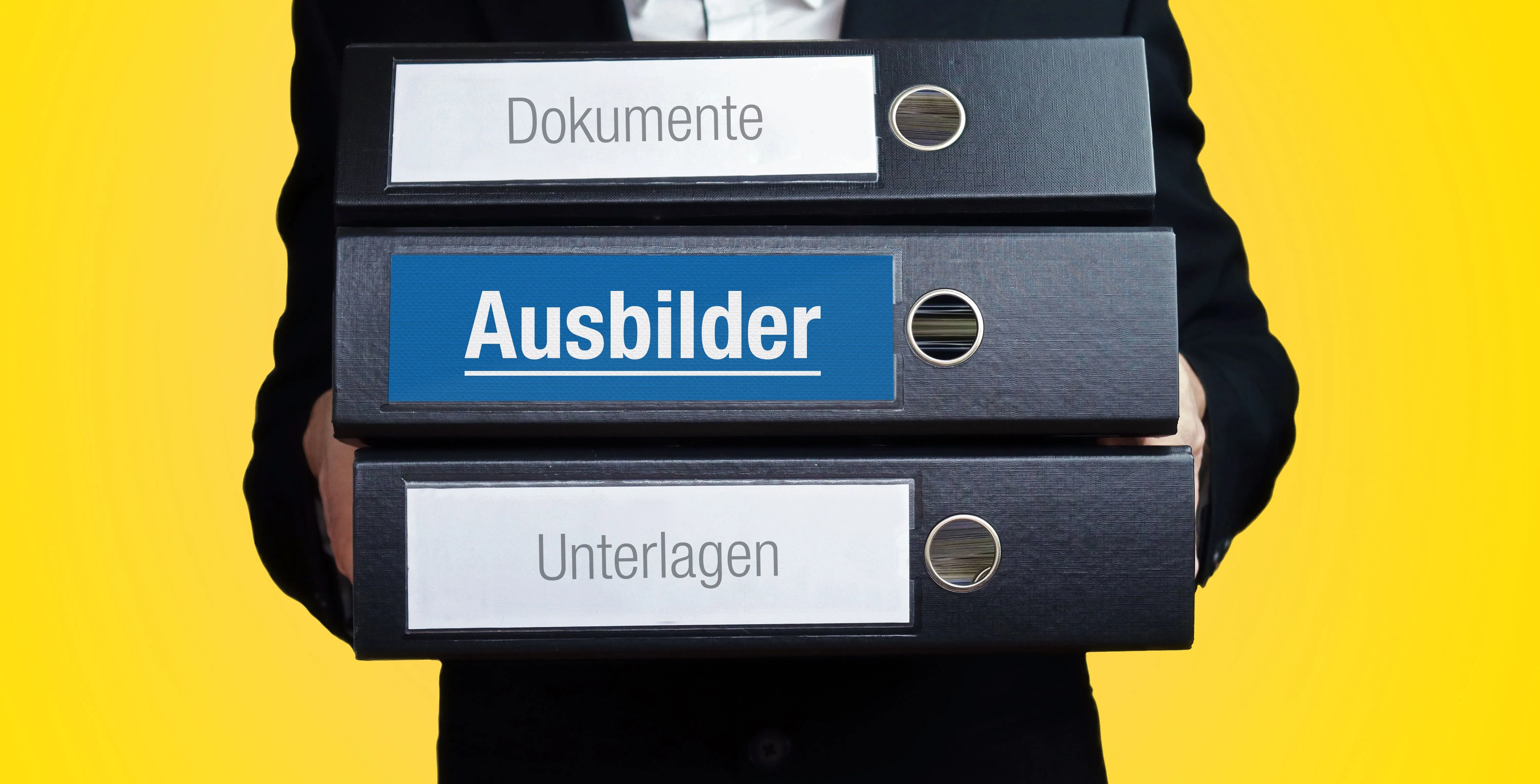 Ein Ausbilder trägt drei Ordner. SIe sind mit folgenden Wörtern beschriftet: Dokumente, Ausbilder, Unterlagen.