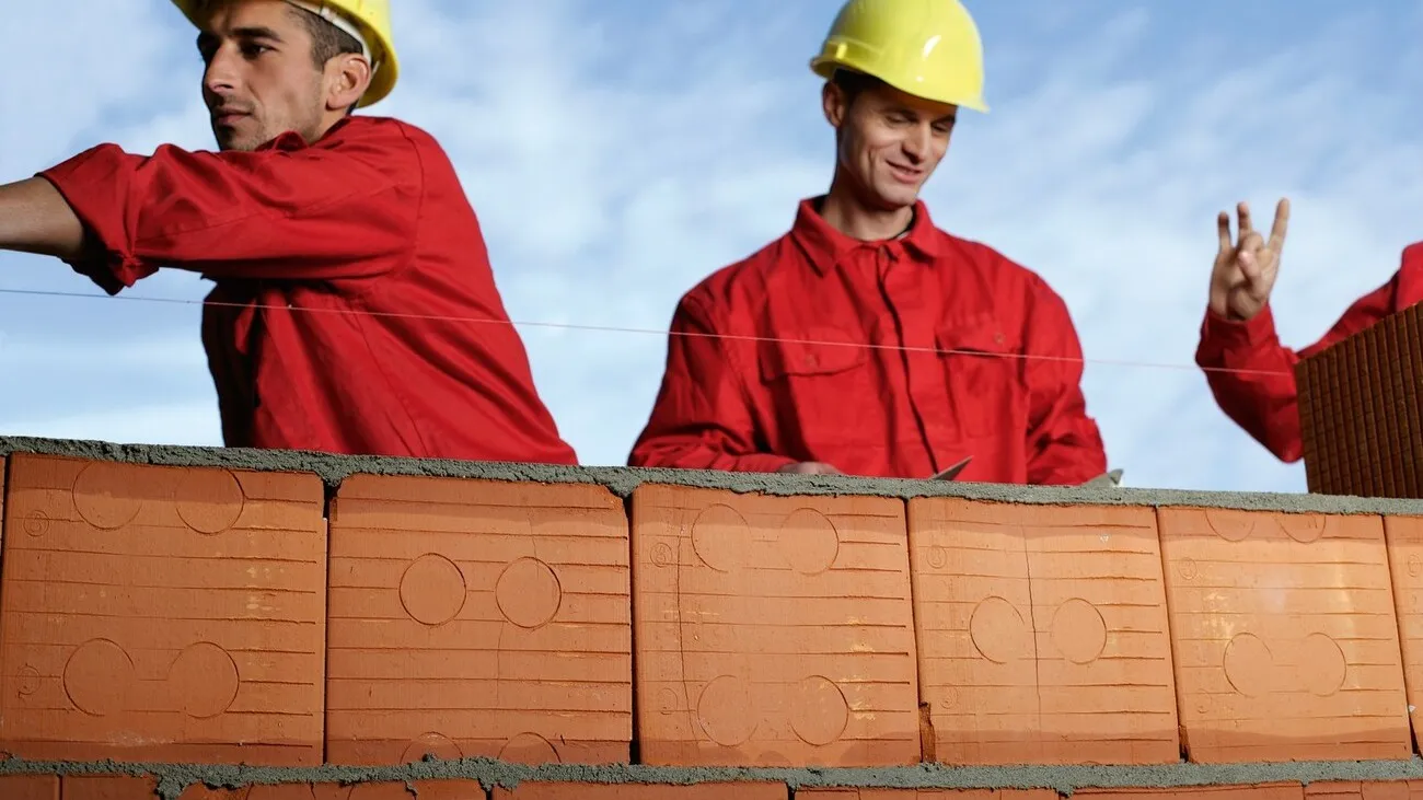 Drei Bauarbeiter mit gelben Schutzhelmen und roter Kleidung arbeiten hinter einer Ziegelmauer.