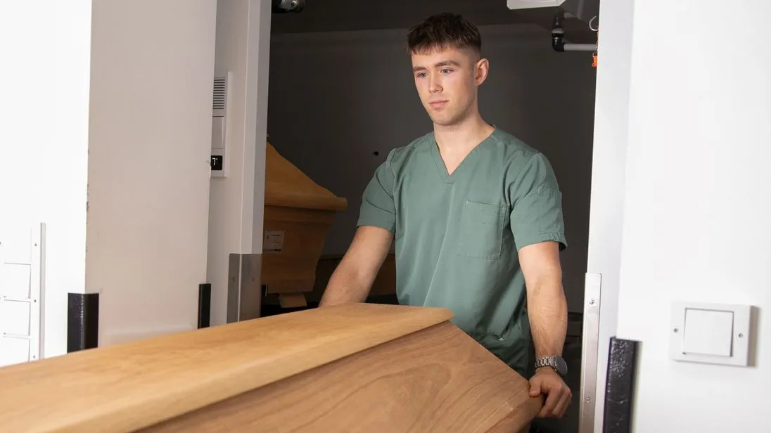 Luis Bauer fährt einen geschlossenen Sarg aus hellbraunem Holz durch eine Tür.