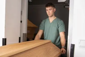 Luis Bauer fährt einen geschlossenen Sarg aus hellbraunem Holz durch eine Tür.