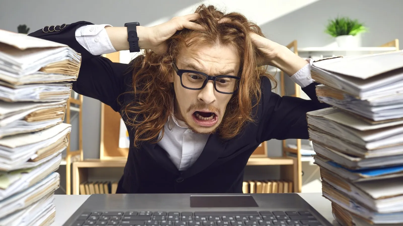 Ein Mann mit langen Haaren und einer schwarzen Brille sitzt vor einem Laptop und hält sich völlig frustriert am Kopf und verzieht das Gesicht.