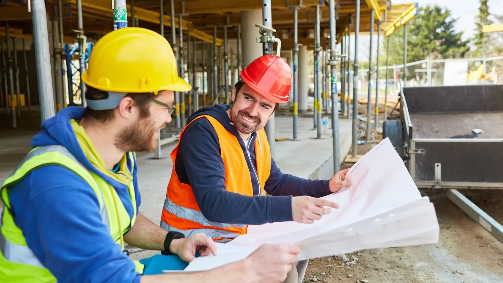Zwei Bauarbeiter sind auf der Baustelle, halten einen Bauplan in den Händen und unterhalten sich.