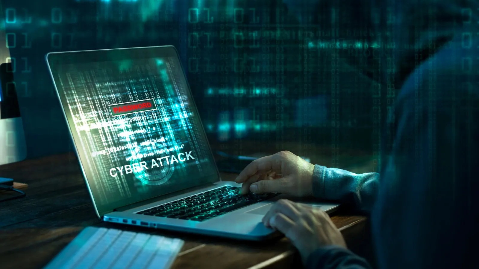 Eine Person mit Kapuze sitzt an einem Laptop. Auf dem Laptopbildschirm ist in Großbuchstaben "Cyber Attack" zu lesen.