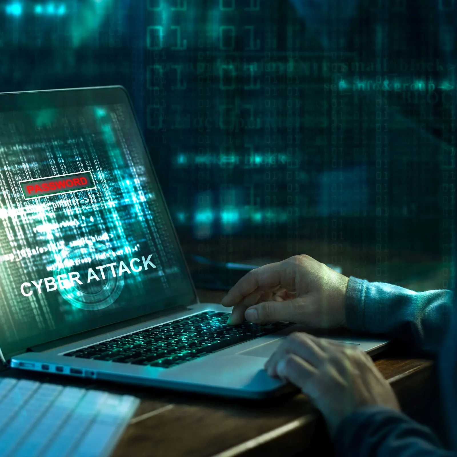 Eine Person mit Kapuze sitzt an einem Laptop. Auf dem Laptopbildschirm ist in Großbuchstaben "Cyber Attack" zu lesen.