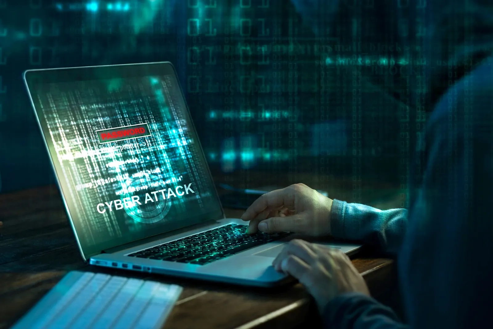 Eine Person mit Kapuze sitzt an einem Laptop. Auf dem Laptopbildschirm ist in Großbuchstaben "Cyber Attack" zu lesen.
