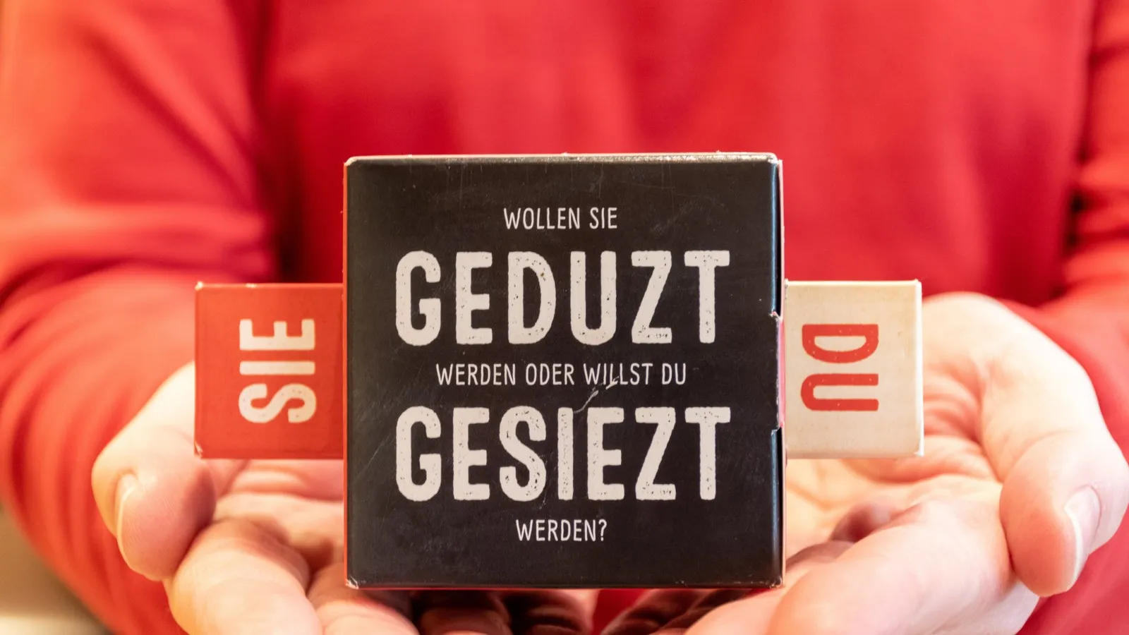 Hände halten einen schwarzen Block mit der Aufschrift "Wollen Sie geduzt werden oder willst du gesiezt werden?", links an diesem Block ist ein rotes "Sie" platziert und rechts ein weißes "Du".
