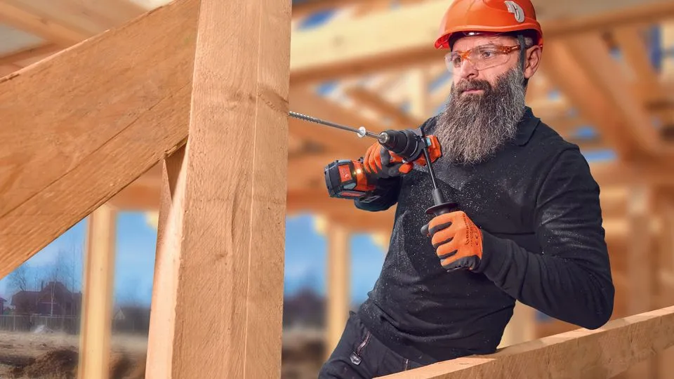 Ein Bauarbeiter mit Bart, Helm und Schutzbrille bohrt in einen Holzbalken.