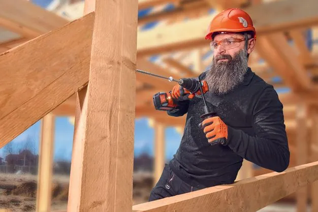 Ein Bauarbeiter mit Bart, Helm und Schutzbrille bohrt in einen Holzbalken.