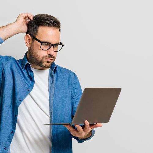 Mann schaut verzweifelt auf sein Laptop, das er in der Hand hält und kratzt sich am Kopf.