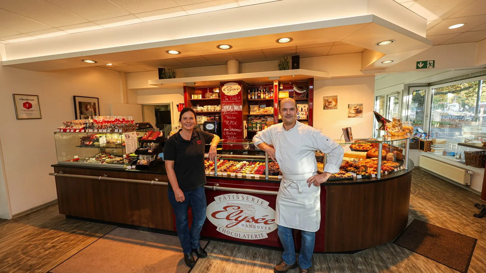Die Gründer der Patisserie Elysee stehen vor einem Tresen voller süßer Leckereien. Links im Bild ist Christina Hochheuser zu sehen. Rechts neben ihr steht der Meisterkonditor Serge Maranzana.