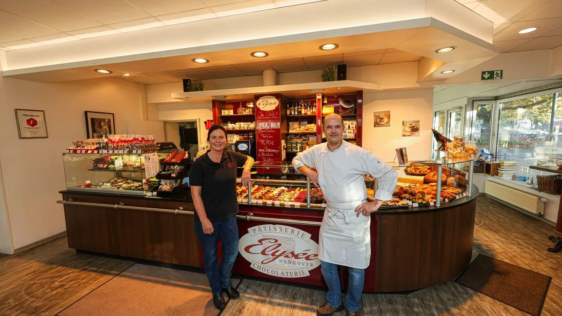Die Gründer der Patisserie Elysee stehen vor einem Tresen voller süßer Leckereien. Links im Bild ist Christina Hochheuser zu sehen. Rechts neben ihr steht der Meisterkonditor Serge Maranzana.