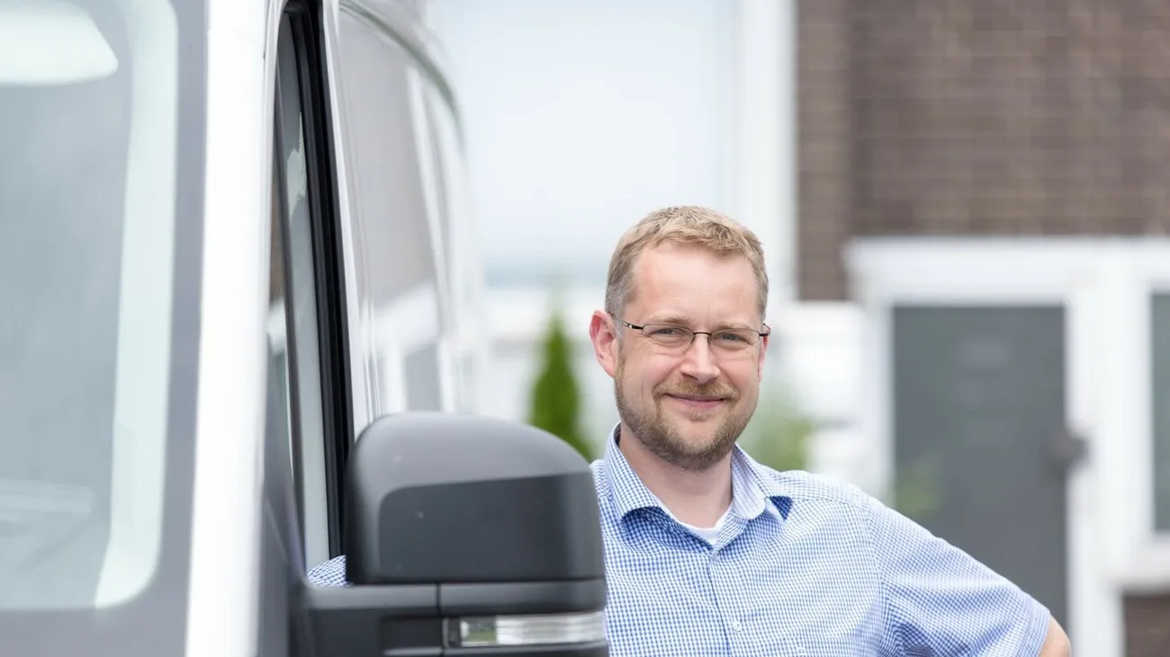 Nahaufnahme eines blonden Mannes mit Brille, der neben einem Transporter steht und lächelt.