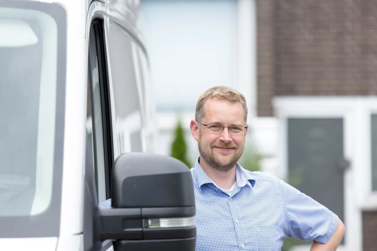 Nahaufnahme eines blonden Mannes mit Brille, der neben einem Transporter steht und lächelt.