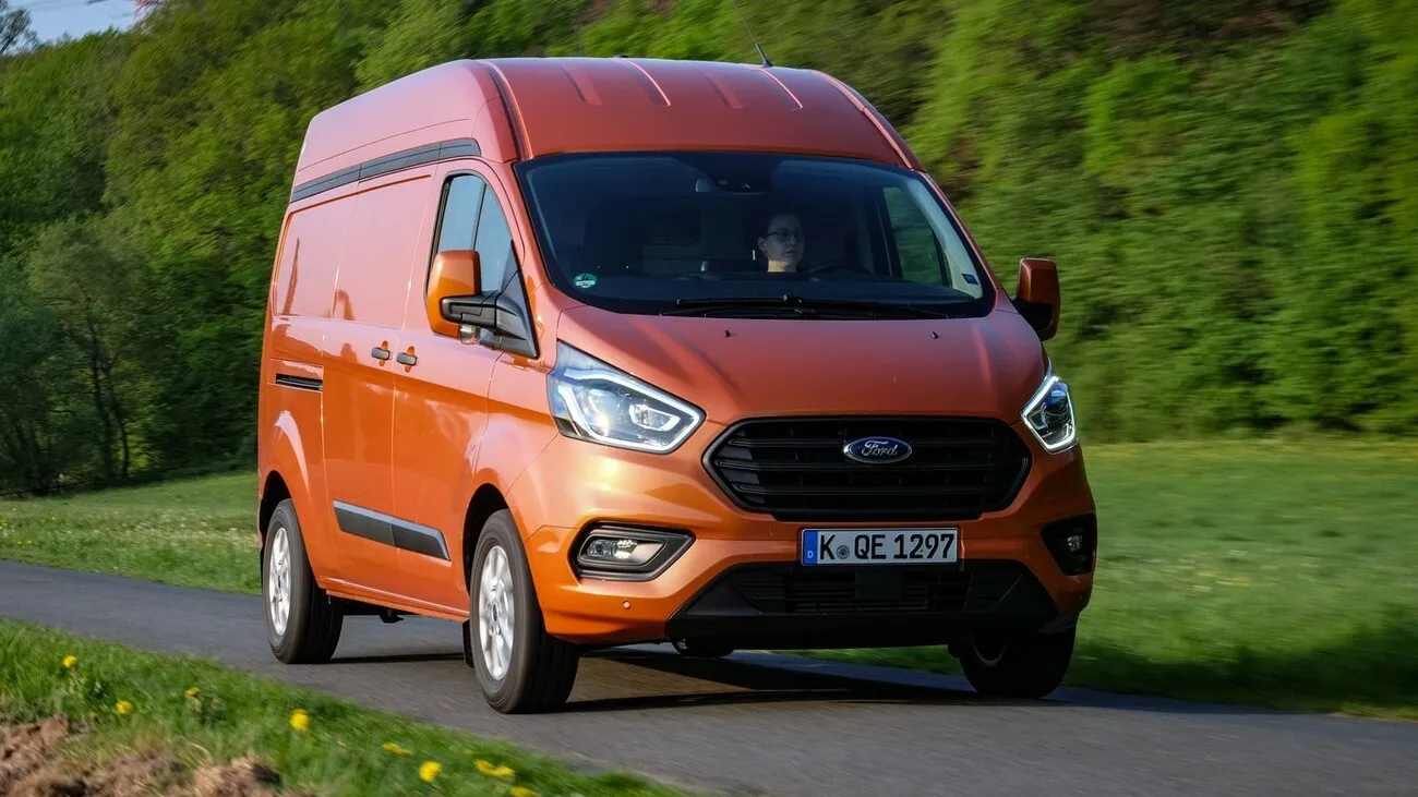 Der Ford Transit Custom in orange steht auf einer Straße, dahinter sind Bäume.