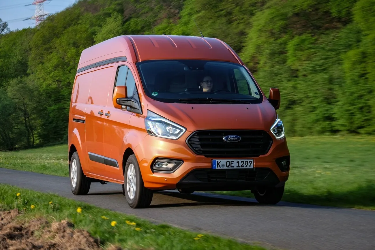 Der Ford Transit Custom in orange steht auf einer Straße, dahinter sind Bäume.