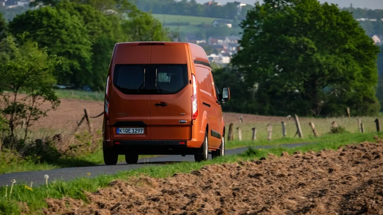 Der orangene Ford Transit Custom von hinten. Er steht auf einer grünen Landschaft mit Feldern im Hintergrund.