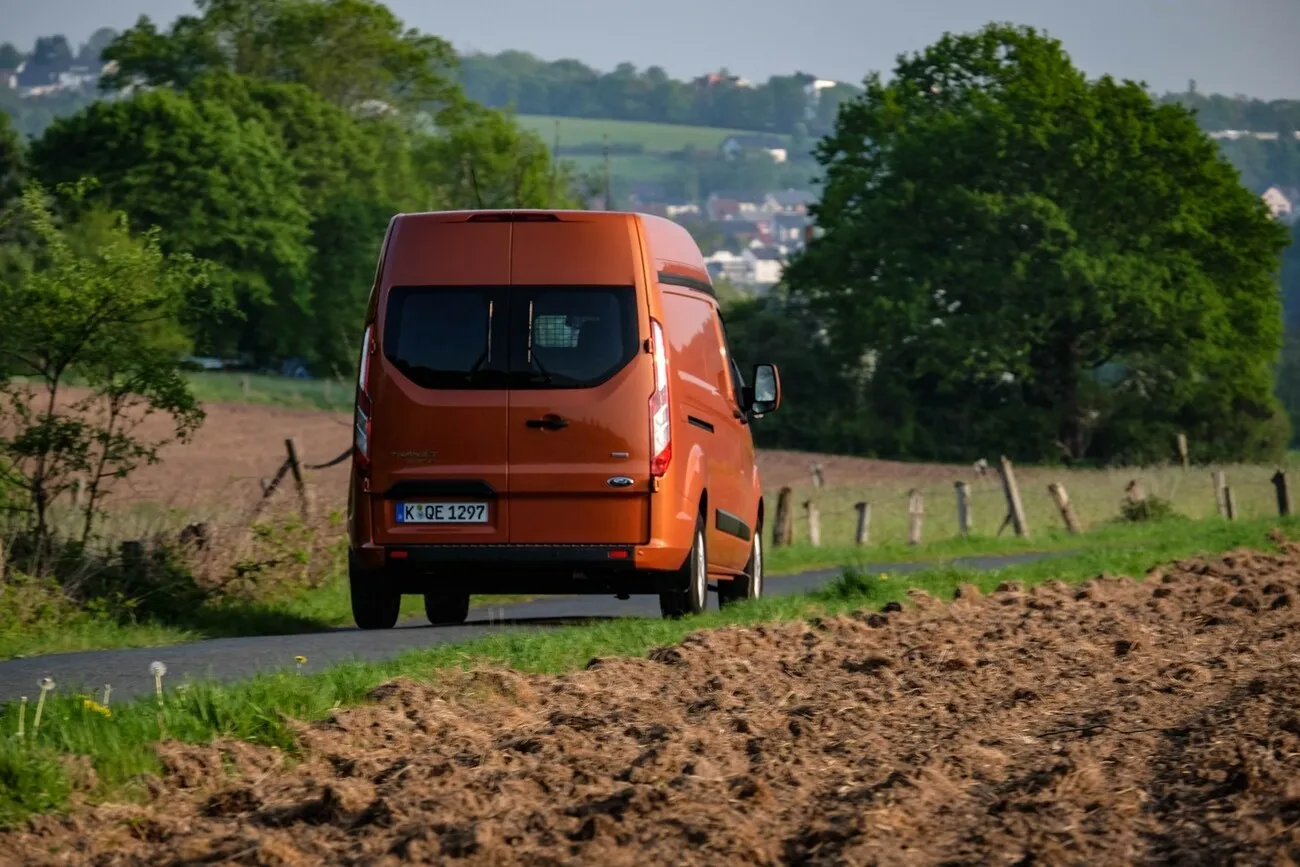 Der orangene Ford Transit Custom von hinten. Er steht auf einer grünen Landschaft mit Feldern im Hintergrund.