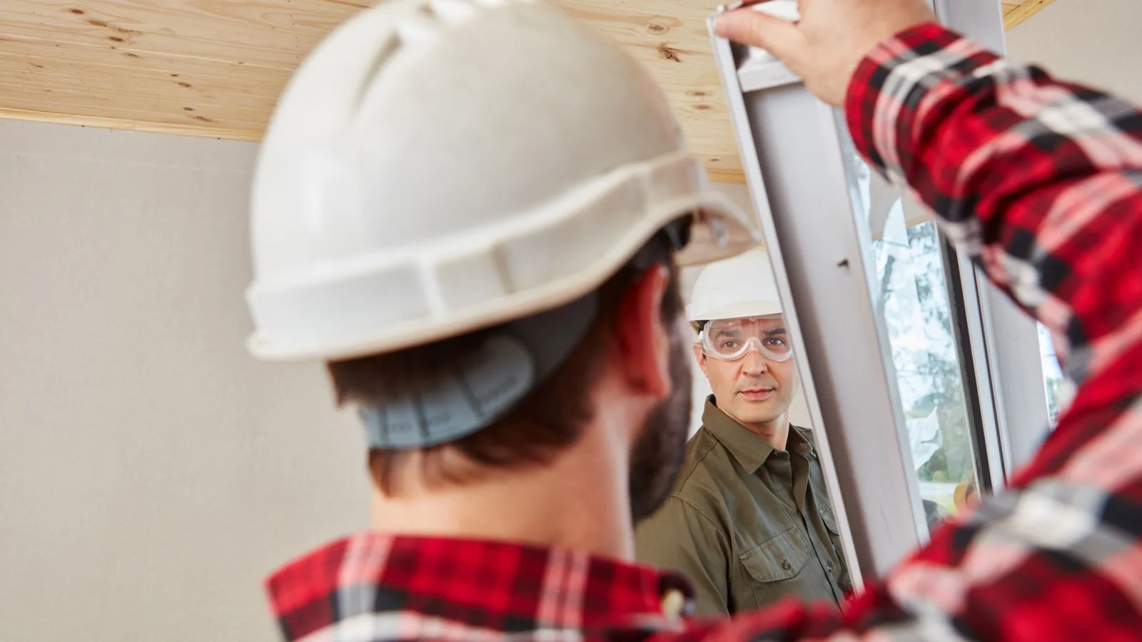 Zwei Handwerker tragen weiße Schutzhelme beim Tragen eines Fensters.