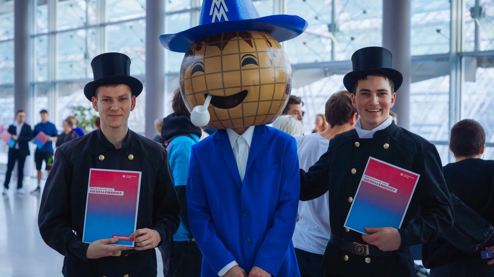 Zwei Männer in traditioneller Schornsteinfegerbekleidung stehen rechts und links von einer Person, die einen braunen Globus auf dem Kopf hat und blau gekleidet ist.
