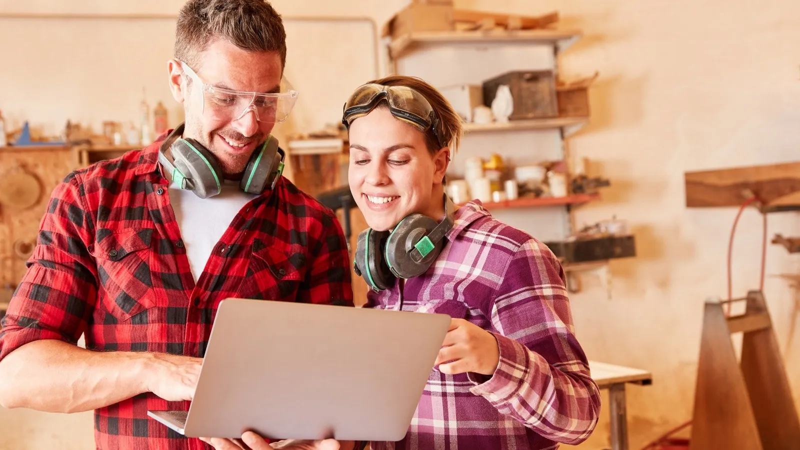 Ein Handwerker und eine Handwerkerin stehen in einer Werkstatt und schauen zusammen auf einen Laptop.