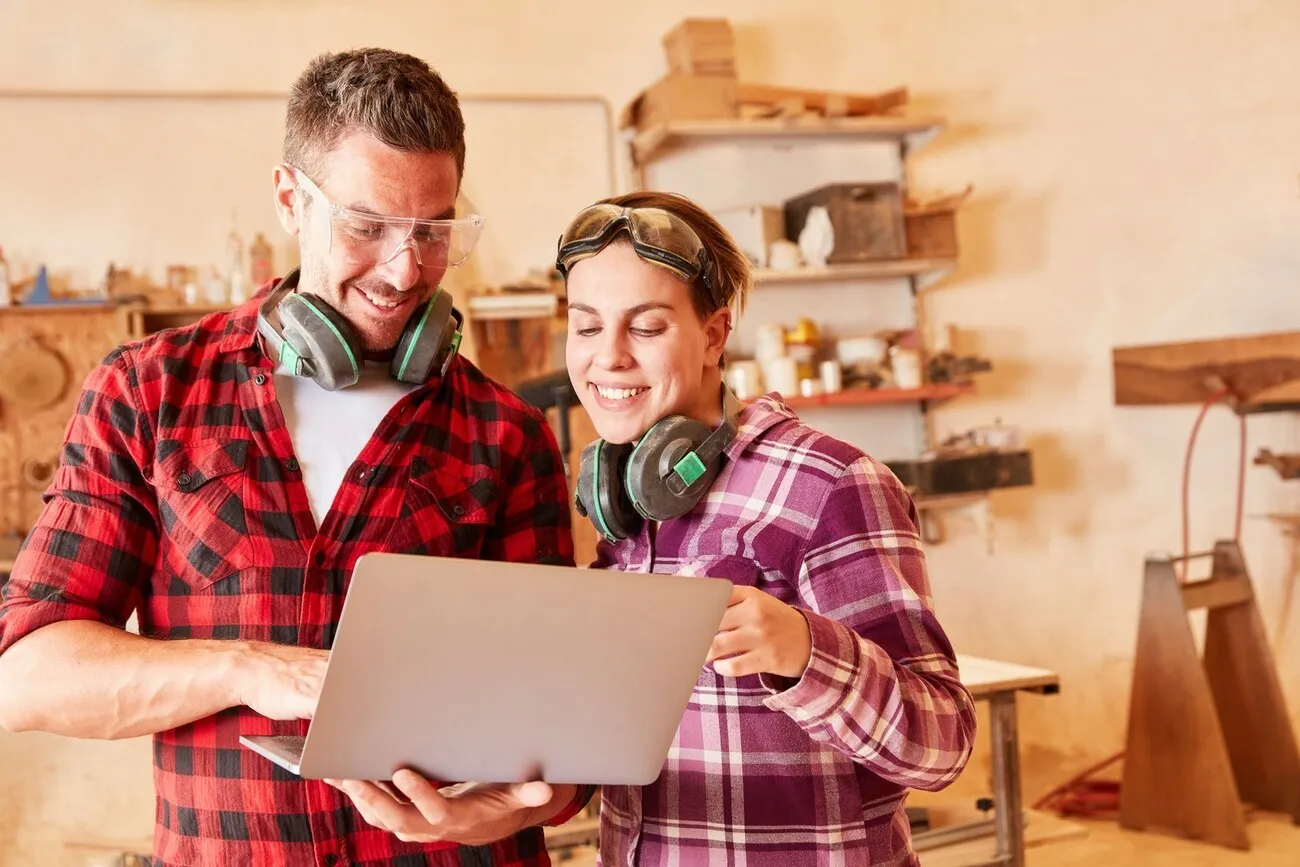 Ein Handwerker und eine Handwerkerin stehen in einer Werkstatt und schauen zusammen auf einen Laptop.