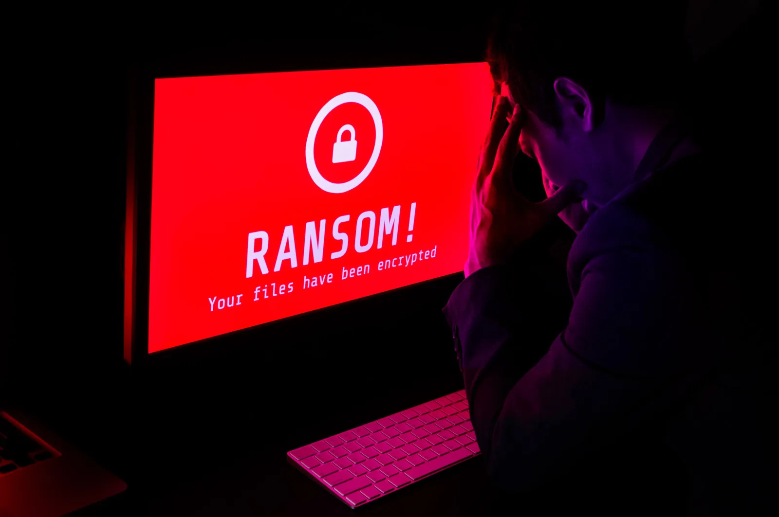 Ein Mann sitzt im Dunkeln vor einem PC-Bildschirm, der rot aufleuchtet und eine weiße Aufschrift "RANSOM" hat.