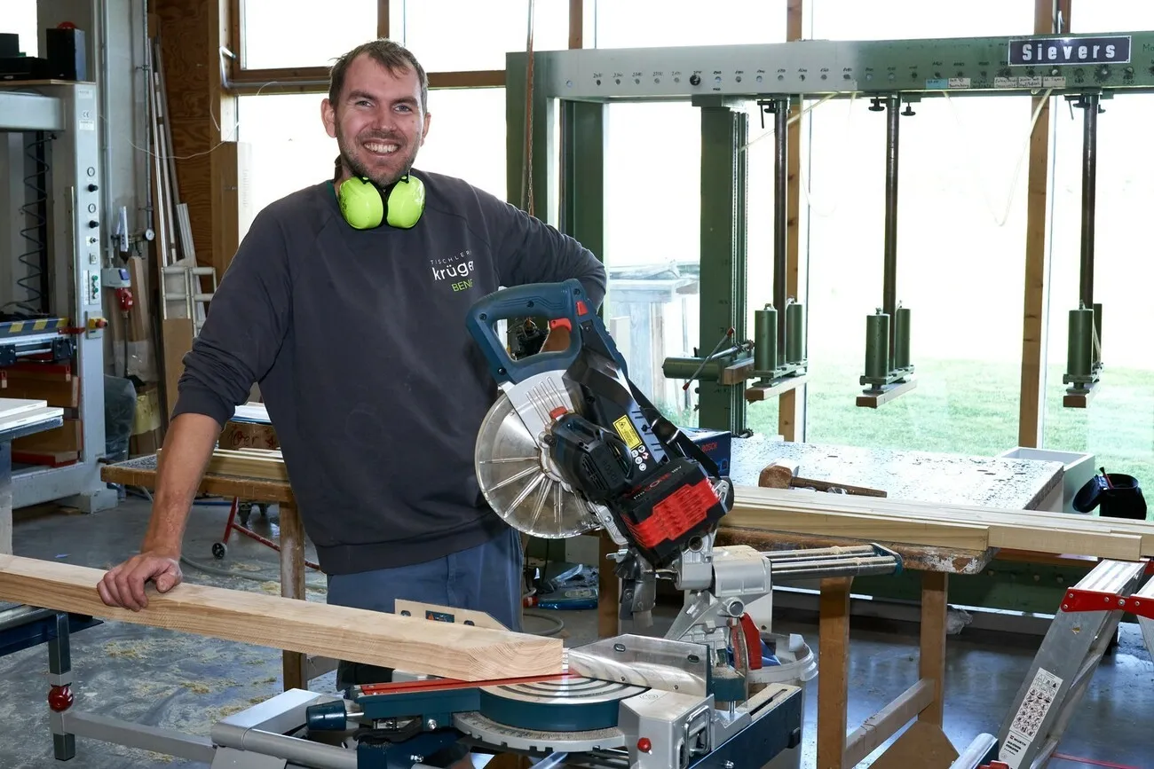 Ein Handwerker steht an der Akku-Klappsäge in einer Werkhalle und lächelt in die Kamera.