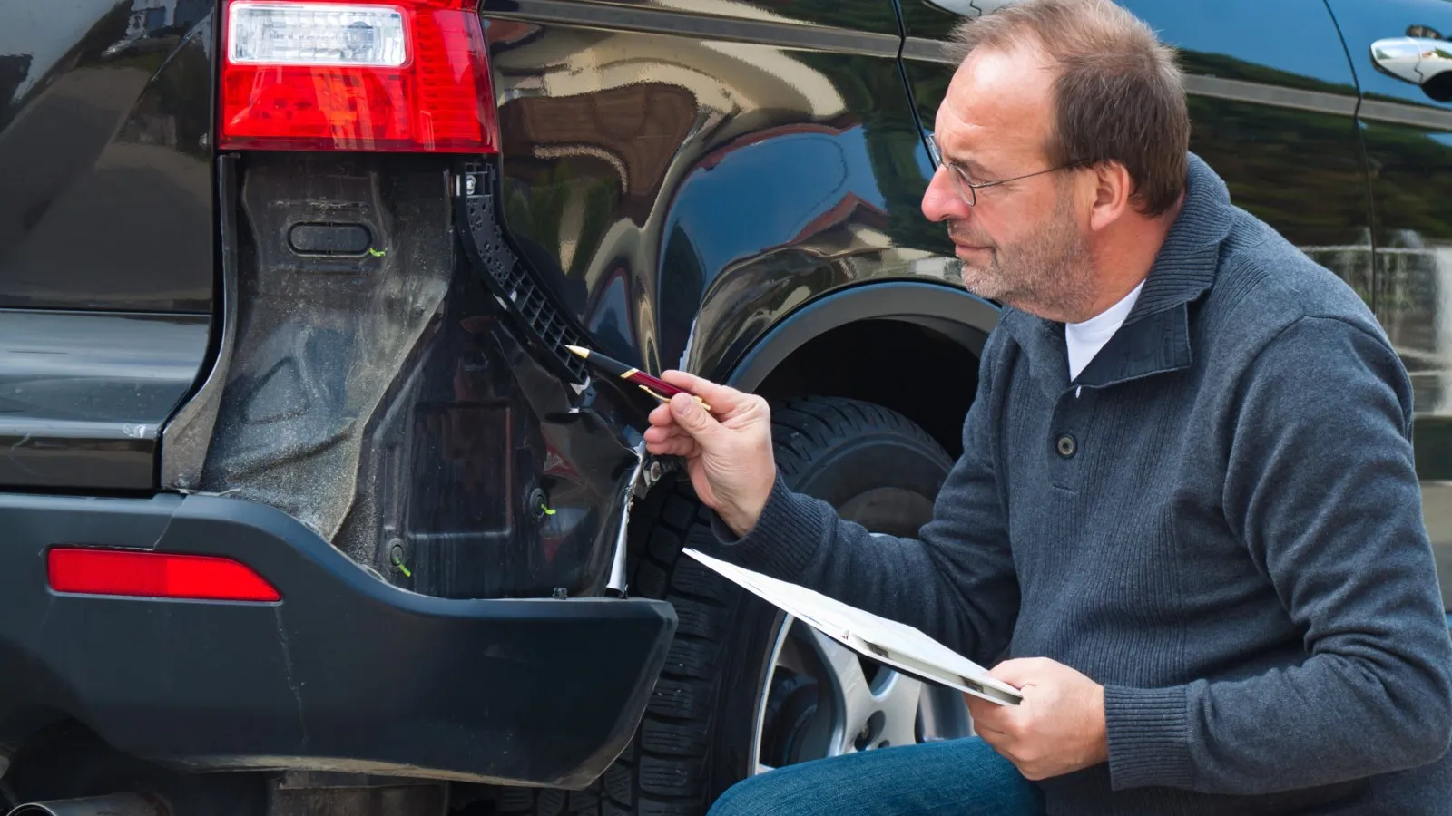 Ein Kfz-Sachverständiger hockt neben einem schwarzen Auto, an dem ein Stück der Karosserie fehlt.