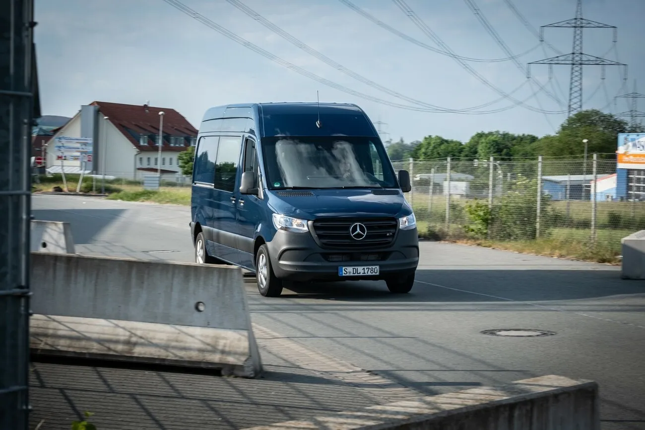 Ein dunkelblauer Mercedes Transporter steht mitten auf einer Straße. Im Hintergrund ist ein Haus und eine Grünfläche zu sehen.